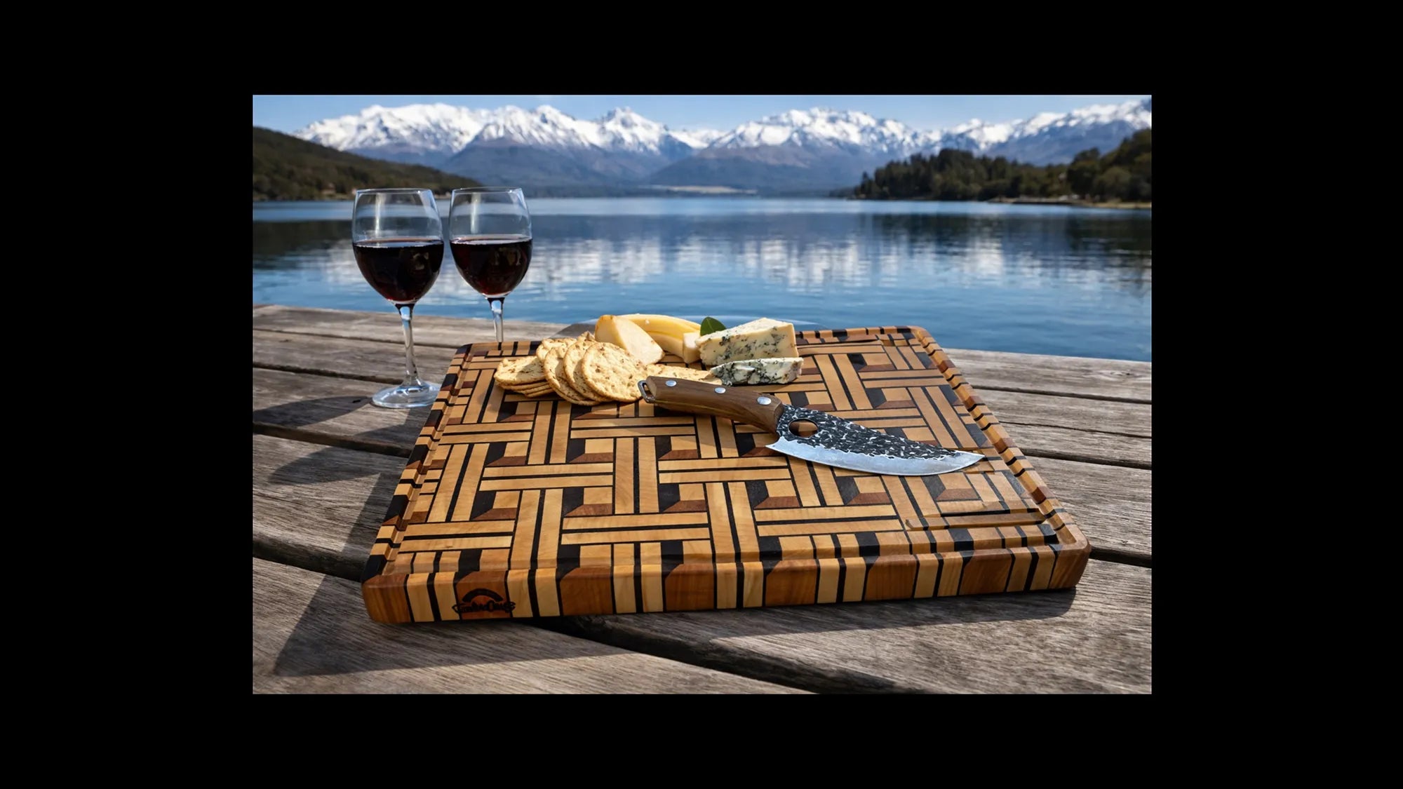 End grain chopping board with cheese and wine glasses on a wooden deck by a lake with snow capped mountains in the background.